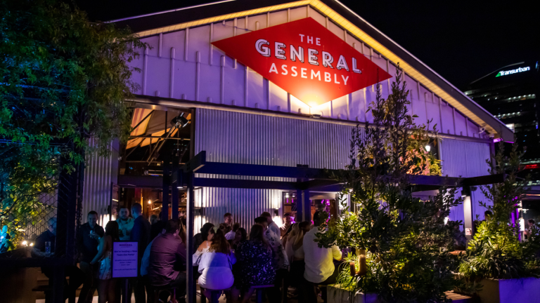 The illuminated sign of The General Assembly stands out against the night sky, surrounded by bustling crowds, highlighting the venue's appeal for evening events and venue hire.